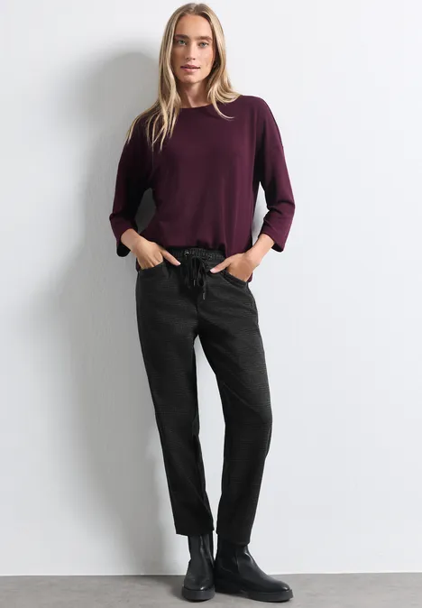 Frau vor weißem Hintergrund in bordeauxfarbenem Pullover, schwarzer Hose und schwarzen Boots.