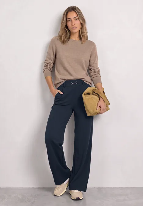 Vrouw in beige trui, wijde donkerblauwe broek en sneakers, met een beige tas in haar hand.