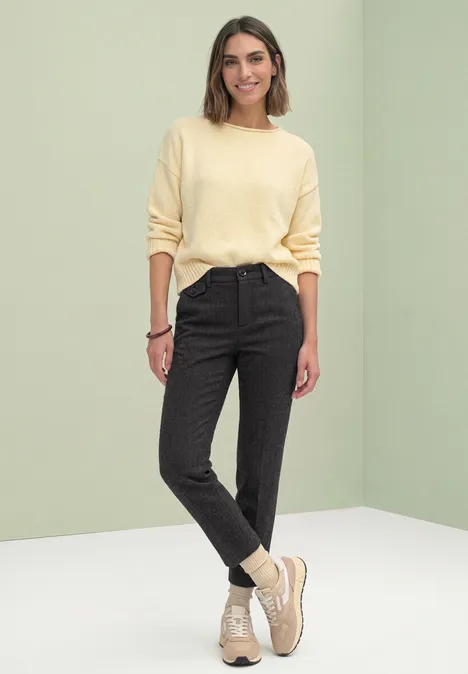 Femme en pull jaune pâle, pantalon gris foncé et baskets beiges sur fond vert clair.