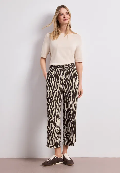 Eine Person trägt ein cremefarbenes Kurzarm-T-Shirt und eine weite zebragemusterte Hose mit braunen Schnürschuhen.