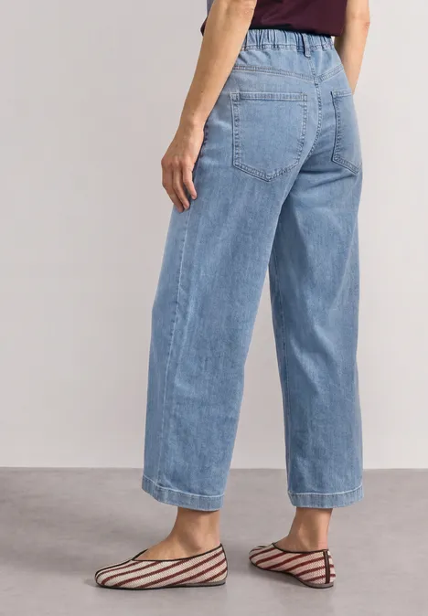 Mid Waist Jeansculotte mit Elasikbund Light Blue Random Washed Frau trägt helle, weite Jeanshose bis Knöchel und flache, gestreifte Ballerina-Sandalen.