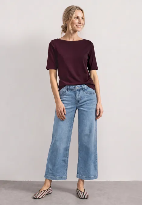Mid Waist Jeansculotte mit Elasikbund Light Blue Random Washed Model in weitem Jeans, burgunderfarbenem Shirt und flachen, gestreiften Sandalen vor neutralem Hintergrund.