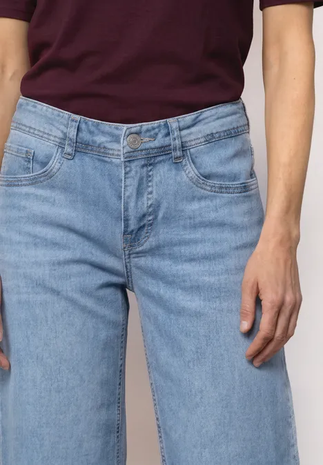 Mid Waist Jeansculotte mit Elasikbund Light Blue Random Washed Eine Person trägt hellblaue Jeans und ein bordeauxfarbenes Oberteil, Fokus auf Taille und Händen.