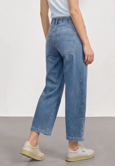 Femme vue de dos portant un jean taille haute ample, baskets claires et haut blanc sur fond neutre.