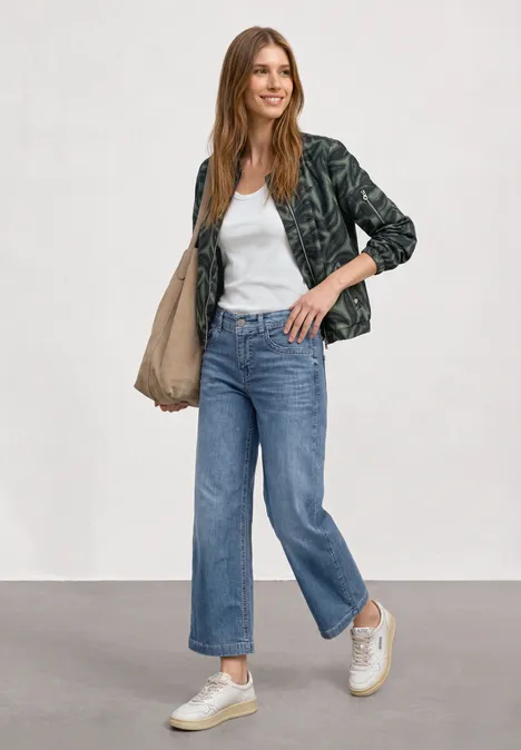 Femme en T-shirt blanc, veste sombre, jean bleu, baskets blanches, sac à la main, fond neutre.