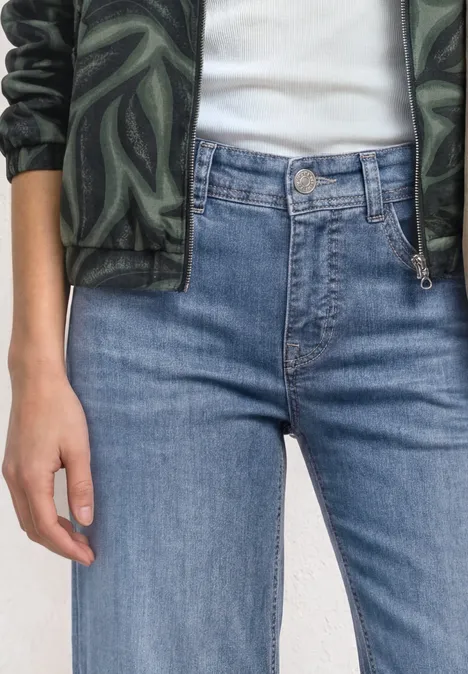 Personne porte un jean taille haute bleu, un t-shirt blanc et une veste verte à motif camouflage.