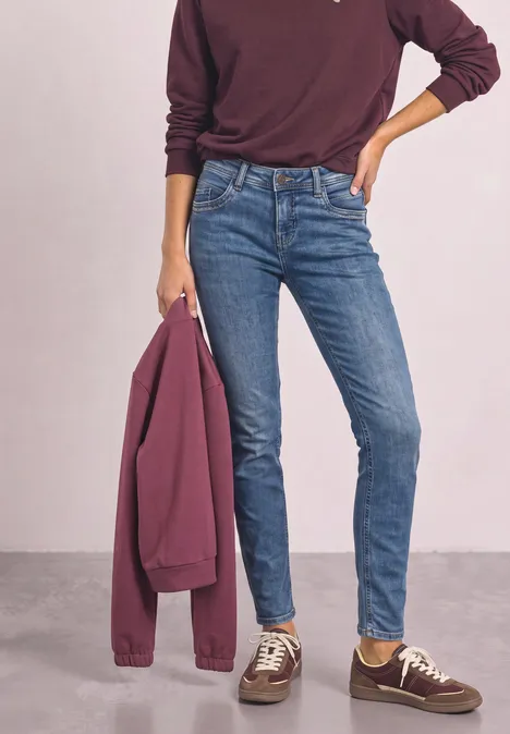 Person trägt schmale blaue Jeans, burgunderfarbenen Pullover, hält passende Jacke und trägt braune Sneakers.