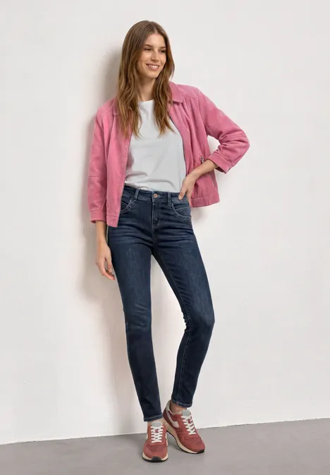 Frau in pinkem Blazer, weißem Shirt, blauen Jeans und roten Sneakern lehnt locker an der Wand.