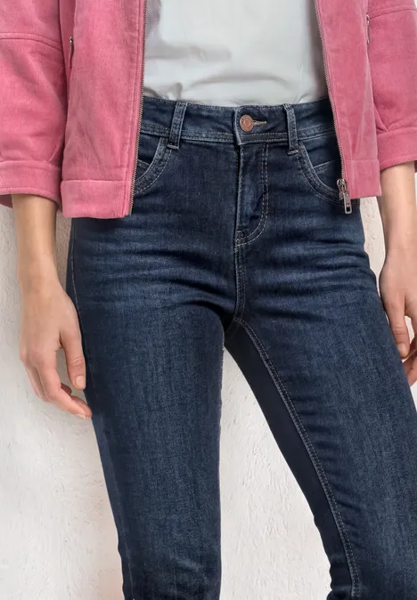 Eine Person trägt dunkelblaue Jeans und eine rosa Jacke, Hände an den Seiten.