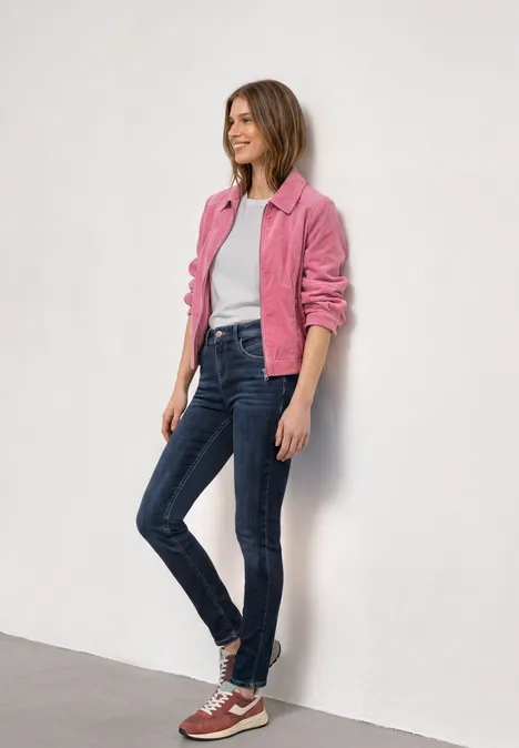 Eine stehende Person vor weißer Wand in pinker Jacke, weißem Shirt, blauen Jeans und roten Sneakern.