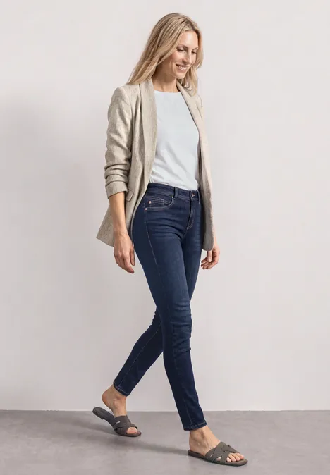 Eine Frau in beige Blazer, weißem T-Shirt, dunkelblauer Jeans und schwarzen Sandalen geht.