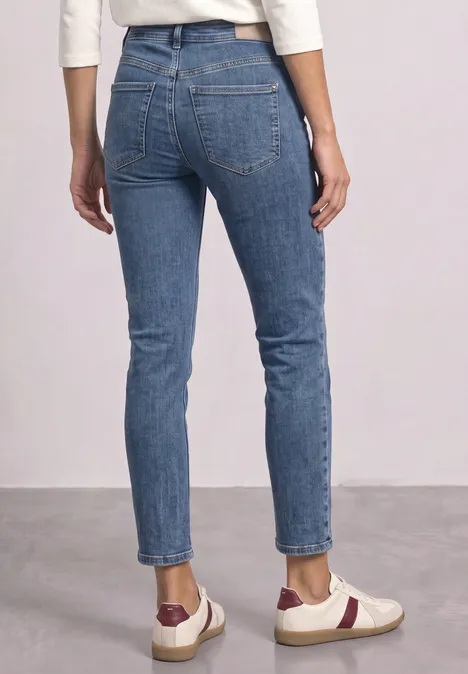 Jeansy Mid Waist z prostymi nogawkami w stylu Slim Fit Medium Blue Soft Washed Osoba od tyłu w dopasowanych niebieskich dżinsach i białych trampkach z czerwonymi dodatkami.
