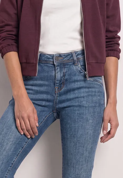 Jeansy Mid Waist z prostymi nogawkami w stylu Slim Fit Medium Blue Soft Washed Osoba w białym T-shircie, bordowej bluzie z kapturem i niebieskich dżinsach z wysokim stanem.