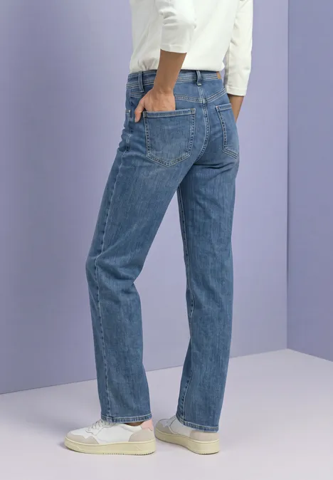 Jeansy Mid Waist z prostymi nogawkami w stylu Casual Fit Medium Blue Soft Washed Widok od tyłu: osoba w prostych niebieskich dżinsach i białych trampkach na fioletowo-szarym tle.