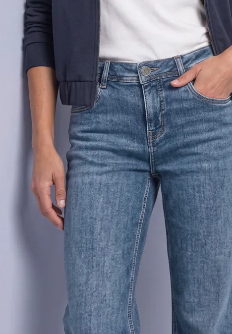Jeansy Mid Waist z prostymi nogawkami w stylu Casual Fit Medium Blue Soft Washed Kobieta w niebieskich dżinsach, białej koszulce i ciemnej kurtce, ręka w kieszeni
