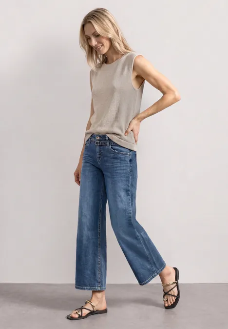 Frau in grauem ärmellosem Top, weiten blauen Jeans und schwarzen Sandalen vor schlichtem grauem Hintergrund.