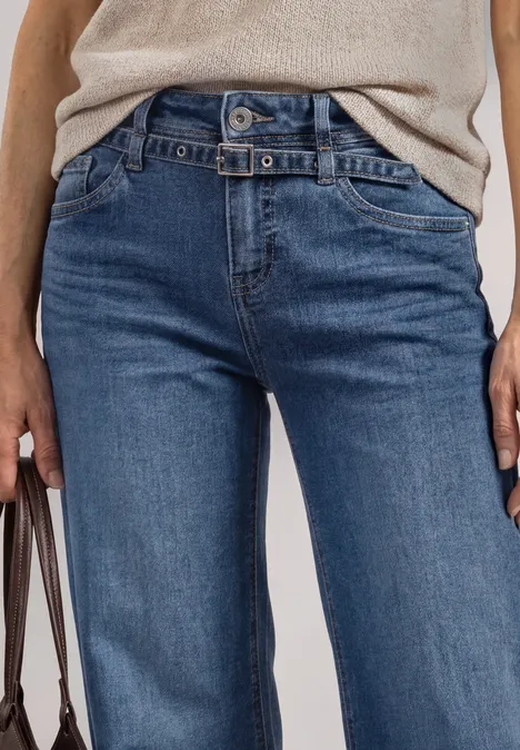 Eine Person trägt eine beige Bluse und blaue Jeans, hält eine Tasche in der linken Hand.