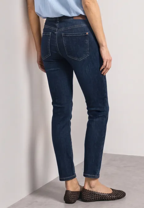 Rückenansicht einer Frau in dunkelblauen Jeans und flachen, schwarzen Geflecht-Slippern vor einer weißen Wand.