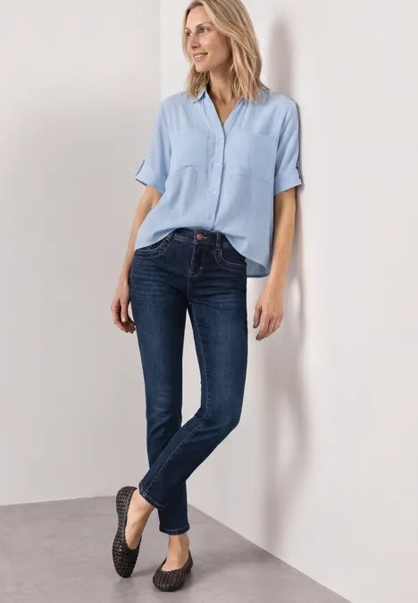Frau lehnt an weißer Wand und trägt hellblaue Bluse, dunkle Jeans und schwarze Ballerinas.