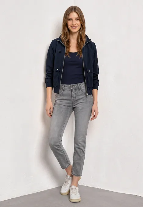 Frau mit dunkelblauer Jacke, schwarzem Shirt, grauen Jeans und weißen Sneakern vor weißem Hintergrund.
