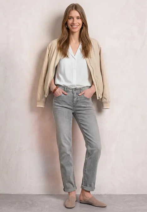 Mid Waist Straight Leg Jeans im Casual Fit Light Grey Random Washed Frau in weißer Bluse, grauer Cordhose und beiger Jacke steht entspannt mit Händen in Taschen vor hellem Hintergrund.