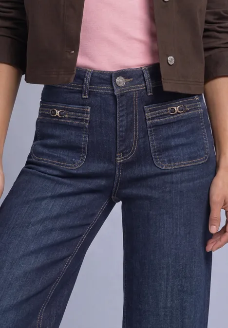 Nahe Aufnahme von hoch sitzenden blauen Jeans mit aufgesetzten Taschen, dazu rosa Top und braune Jacke.