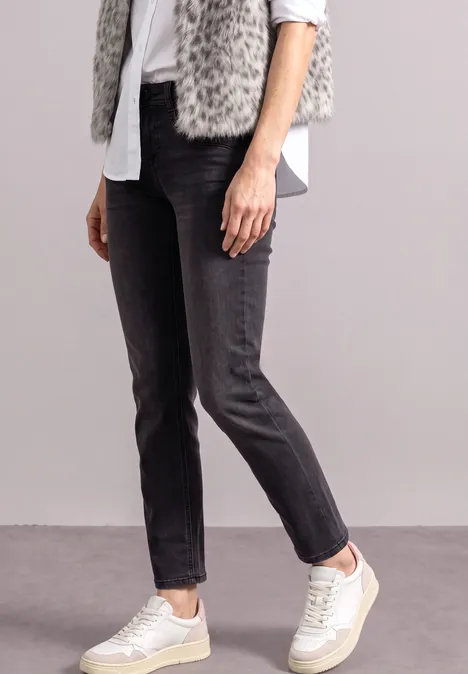 Mid Waist Straight Leg Jeans im Slim Fit Black Washed Person steht in schwarzen Jeans, weißer Bluse, grauer Fellweste und weißen Sneakern vor schlichtem Hintergrund.