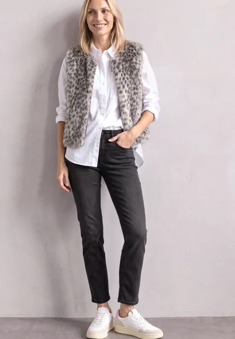 Mid Waist Straight Leg Jeans im Slim Fit Black Washed Frau in weißem Hemd, schwarzer Hose, grauer Fellweste und weißen Sneakers vor hellgrauer Wand.