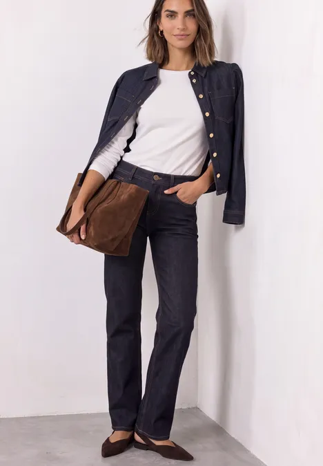 Frau in weißem T-Shirt, dunkelblauem Jackett und Hose, mit brauner Tasche lehnt an weißer Wand.