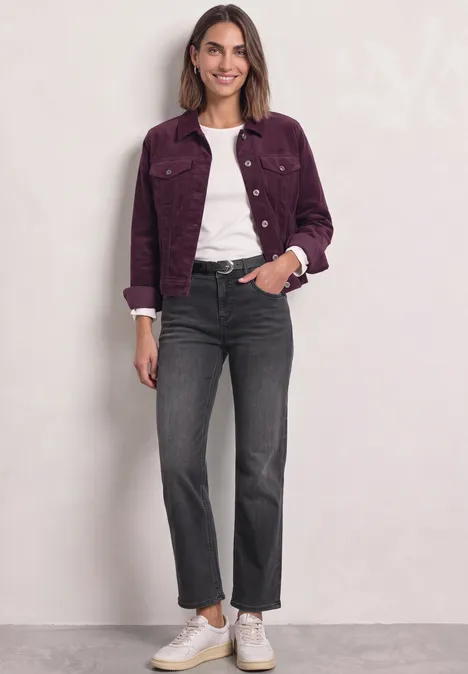 Frau in fliederfarbener Jacke, weißem Shirt, schwarzen Jeans und weißen Sneakern vor hellem Hintergrund.