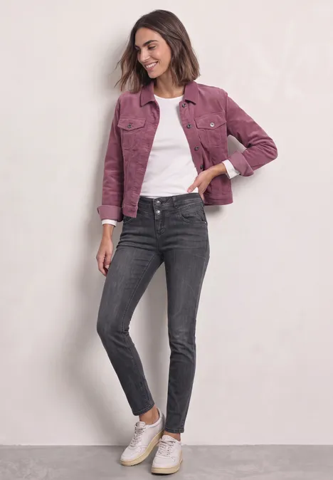 Jonge vrouw met roze jasje, wit T-shirt, donkergrijze jeans en witte sneakers voor een lichte achtergrond.