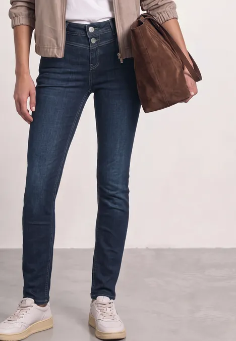 Een persoon draagt donkerblauwe skinny jeans, witte sneakers, een beige jasje en houdt een bruine tas vast.