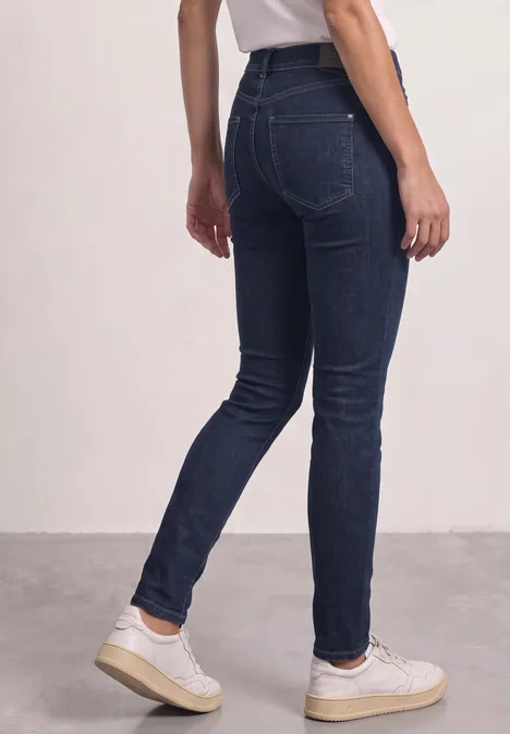 Achteraanzicht van een persoon in donkerblauwe skinny jeans en witte sneakers tegen neutrale achtergrond.