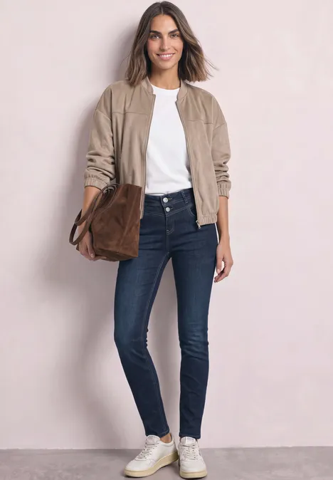 Stijlvolle vrouw in jeans, wit shirt, beige jasje, witte sneakers en bruine tas tegen een lichte achtergrond.