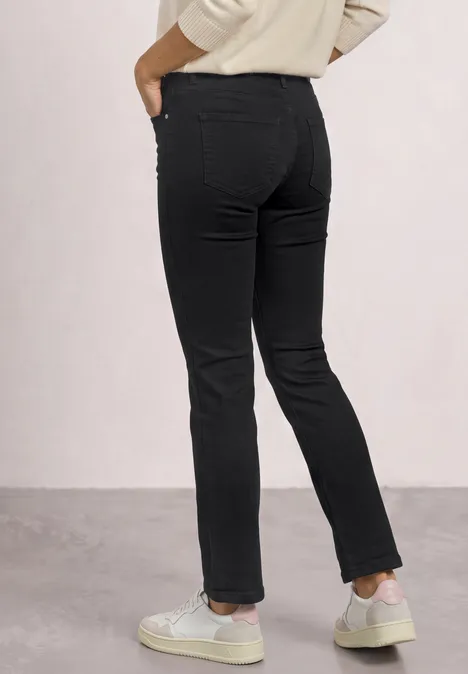 Mid Waist Slim Leg Jeans im Casual Fit Black Clean Washed Frau trägt schwarze, schlanke Jeans und weiße Sneaker, Rückansicht vor hellem Hintergrund.