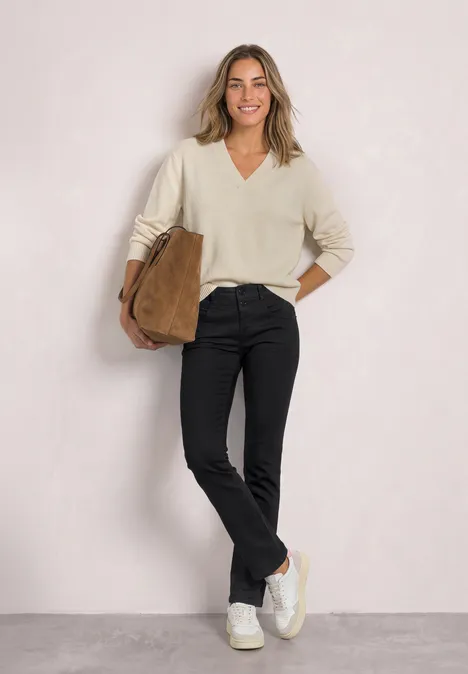 Mid Waist Slim Leg Jeans im Casual Fit Black Clean Washed Eine lächelnde Frau trägt einen cremefarbenen Pullover, schwarze Hose, weiße Sneakers und hält eine braune Tasche.