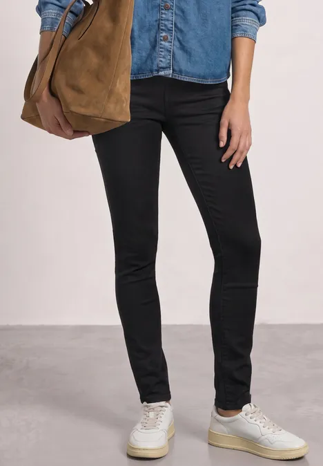 Mid Waist Slim Leg jeans im Slim Fit Black Clean Washed Person in schwarzer Skinny-Jeans, Jeanshemd, weiße Sneaker, trägt olivfarbene Tasche.
