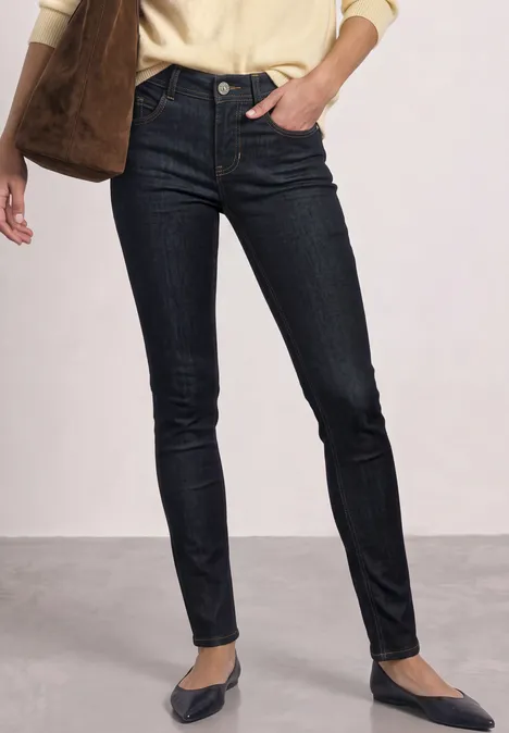 Mid Waist Slim Leg jeans w kroju Slim Fit Deep Rinse Washed Kobieta w ciemnych rurkach i spiczastych balerinach, beżowa bluzka i torebka na ramię.