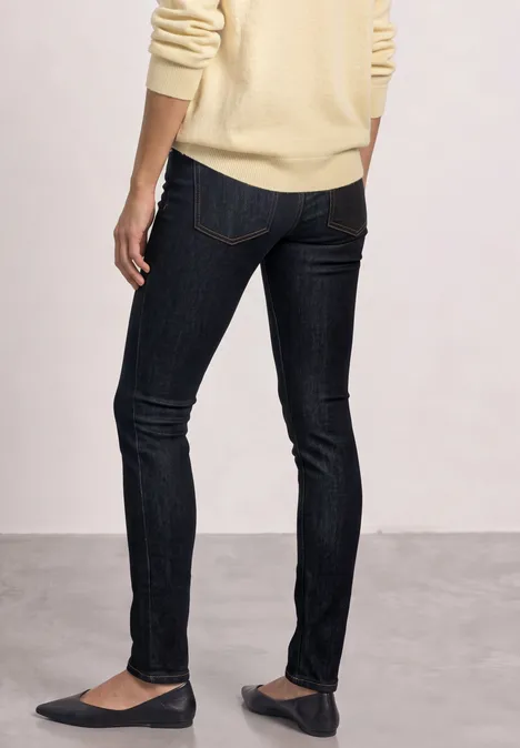 Mid Waist Slim Leg jeans w kroju Slim Fit Deep Rinse Washed Osoba od tyłu w ciemnych dopasowanych dżinsach, beżowym swetrze i balerinach.