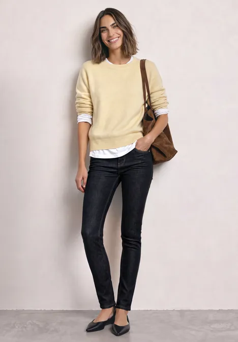 Mid Waist Slim Leg jeans w kroju Slim Fit Deep Rinse Washed Kobieta ma jasny żółty sweter na białej koszuli, czarne jeansy, baleriny i brązową torbę na ramię.