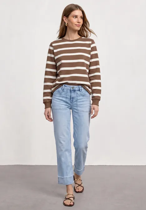 Frau in braun-weiß gestreiftem Pullover, blauen Jeans und Sandalen vor hellem Hintergrund.