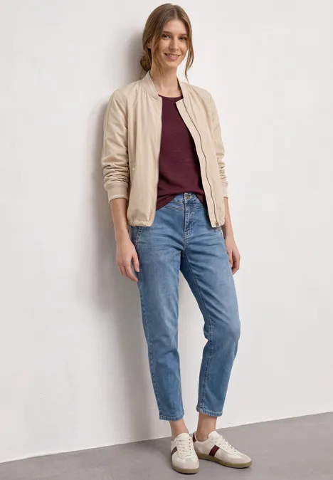 High Waist Barrel Leg Jeans im Loose Fit Medium Blue Random Wash Person trägt beige Bomberjacke, bordeauxfarbenes T-Shirt, blaue Jeans und weiße Sneaker.