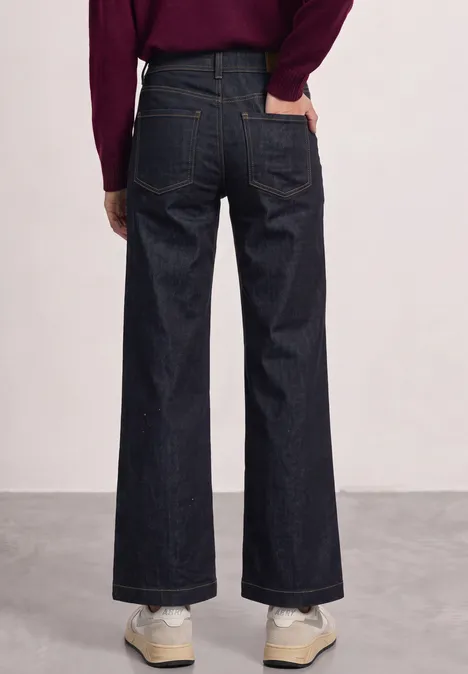 Achteraanzicht van iemand in wijde, donkere jeans en sneakers tegen een effen achtergrond.