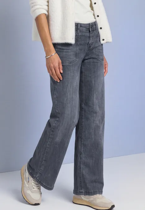 Onderlichaam van persoon in wijde grijze jeans en witte sneakers tegen blauwe achtergrond.