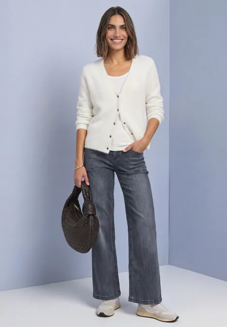 Vrouw in witte cardigan, grijze spijkerbroek, sneakers en ronde rieten tas.