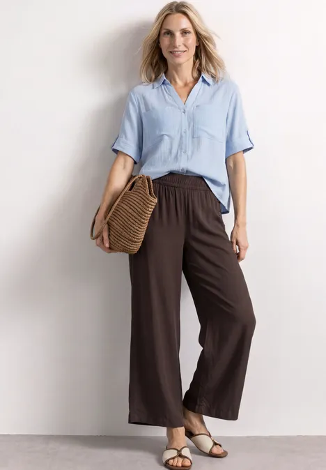 Femme en chemise bleu clair et pantalon large marron, avec sac en paille et sandales, devant un mur neutre.