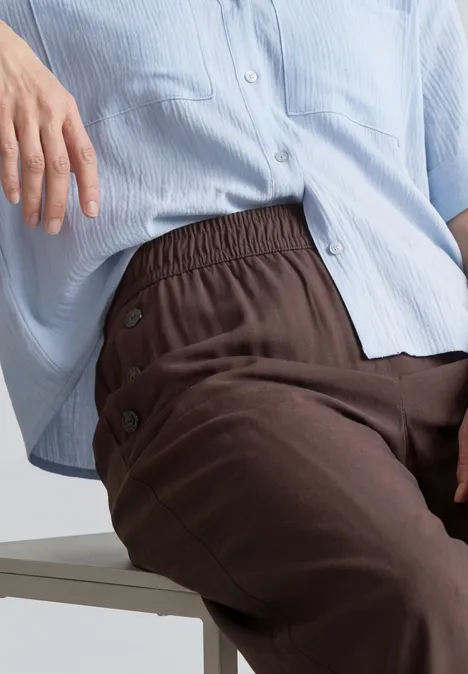 Personne assise portant une chemise bleu clair ample et un pantalon marron foncé à boutons latéraux.
