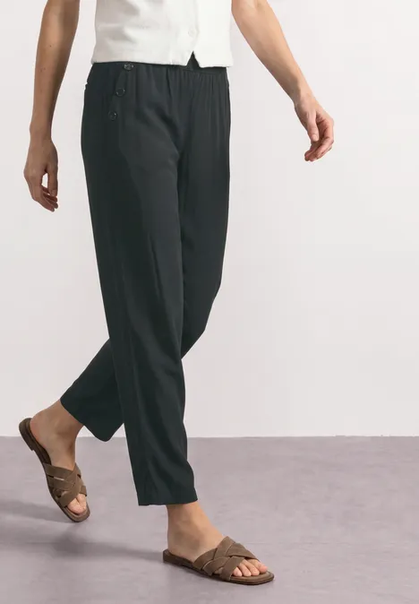 Eine Person trägt schwarze, locker sitzende Hosen und braune Sandalen auf grauem Boden.