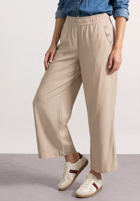 Dame draagt een beige wijdvallende broek met elastische taille en knopen, gecombineerd met witte sneakers met strepen.