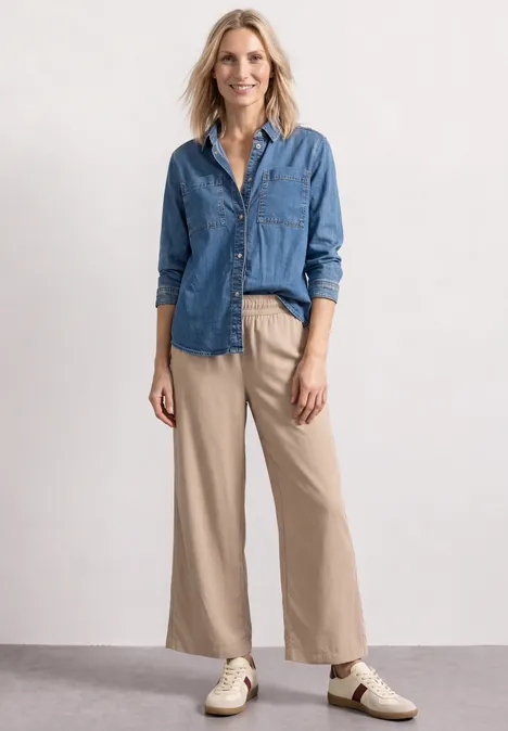 Een vrouw draagt een blauw denim overhemd, beige wijde broek en sneakers, poseert nonchalant.